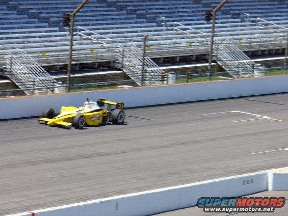 Yellow and white open-wheel race car on a track with empty grandstands in the background