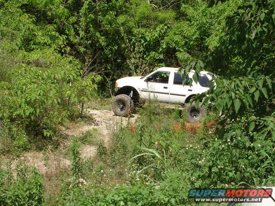 White SUV with large off-road tires and lifted suspension driving on a dirt path surrounded by trees