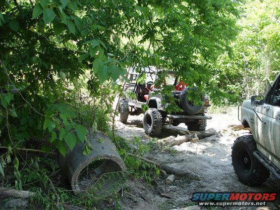 Two off-road vehicles, one a Jeep-like vehicle with a spare tire on the back, the other a truck, on a dirt path surrounded by trees