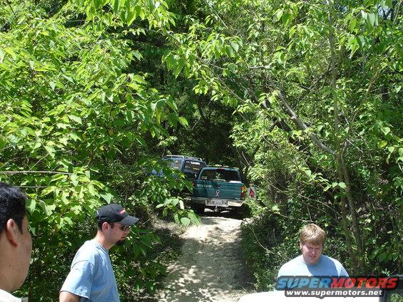 Two trucks driving on a dirt path through trees. People are visible in the foreground