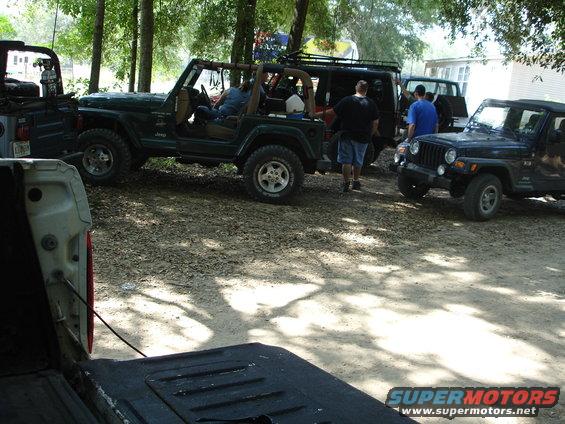 Several Jeeps parked on a dirt lot under trees, with a white pickup truck bed in the foreground