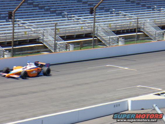 IndyCar race car with orange and white livery on a track, grandstands in background