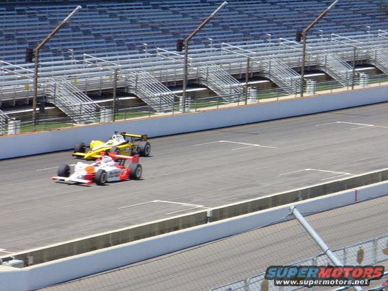 Two open-wheel race cars, one red and white, the other yellow, on a race track with empty grandstands in the background