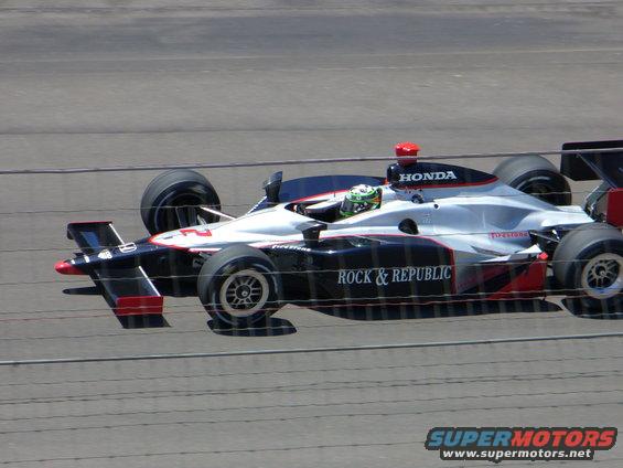 Silver and black IndyCar with red accents, number 2, driven by a person in a green helmet