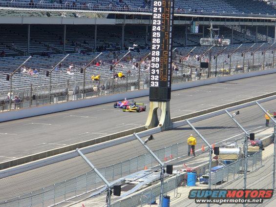 Two IndyCars on a race track with a scoring tower displaying numbers 20-33