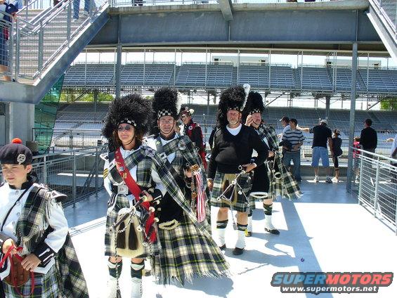 Bagpipe band members in kilts and feathered hats walk past stadium seating