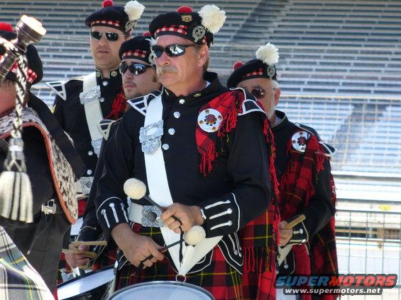 Drummers in kilts and traditional Scottish attire stand in formation, some holding drumsticks