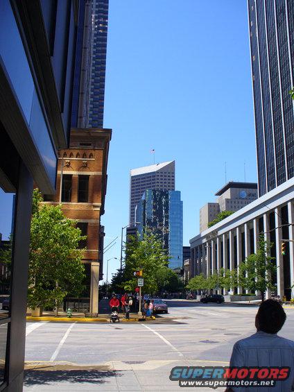 City street with tall buildings, trees, and a few cars visible. A person looks towards the buildings