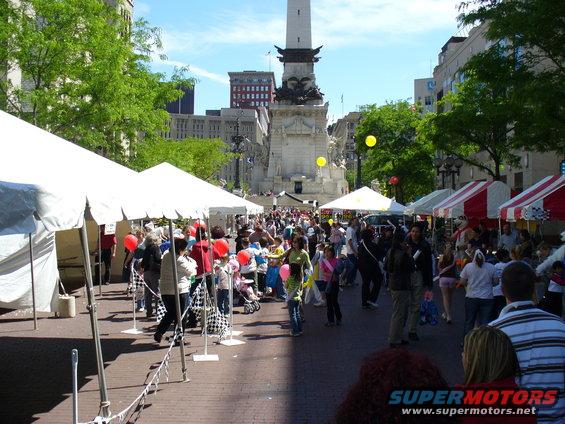 Outdoor festival with tents, crowds, and a tall monument in the background