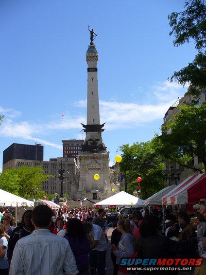 Crowded plaza with a tall monument, tents, and trees under a blue sky