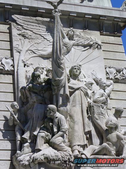 stone sculpture of people holding a flag with an eagle on top, with a tree and sunburst in the background