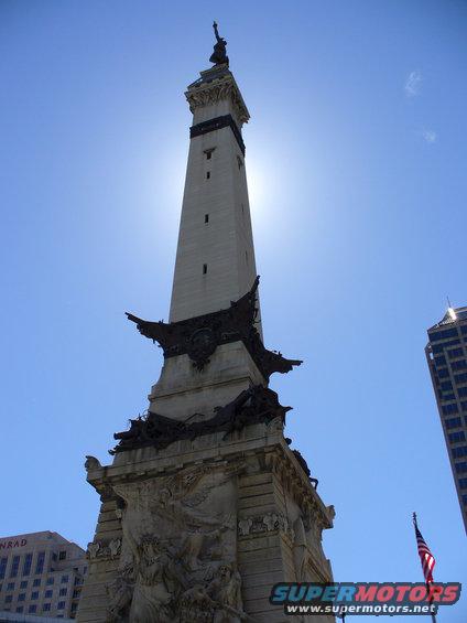 Monument with intricate carvings and a statue on top, against a bright blue sky. A modern skyscraper is visible to the right
