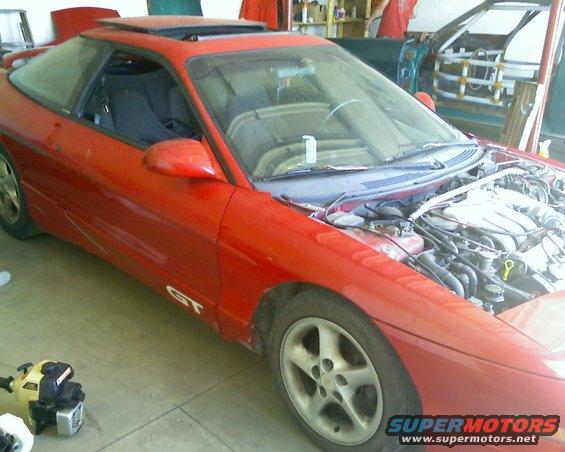 Red 1993 Ford Probe GT with hood open, showing engine bay. Another car body is visible in the background
