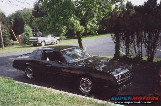 Black coupe parked curbside, 1980s body style. Pickup truck in background
