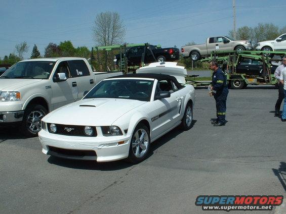 White convertible Ford Mustang with black top, front bumper taped, parked next to a white pickup truck