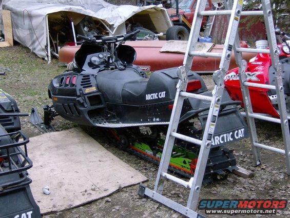 Black Arctic Cat snowmobile parked outside a tent with ladders nearby. Another red snowmobile is partially visible