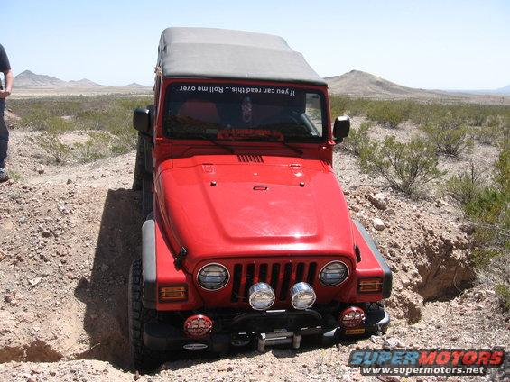 Red Jeep Wrangler on dirt trail, front view. Sticker on windshield says 'If you can read this... Roll me over'