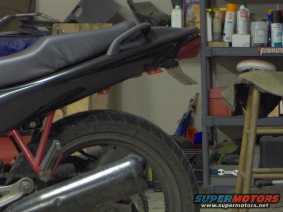 Motorcycle rear seat, tail light, and exhaust pipe visible. Shelving with supplies in background