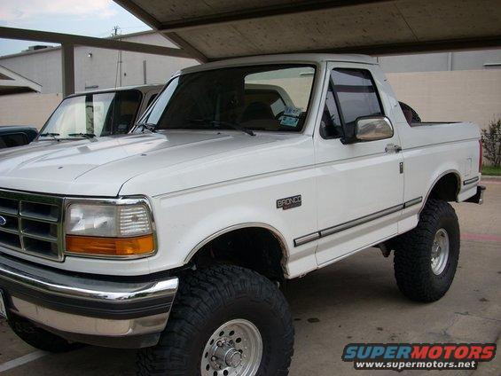 White 1992 Ford Bronco with lifted suspension and large tires parked under a carport. Another vehicle is visible behind it