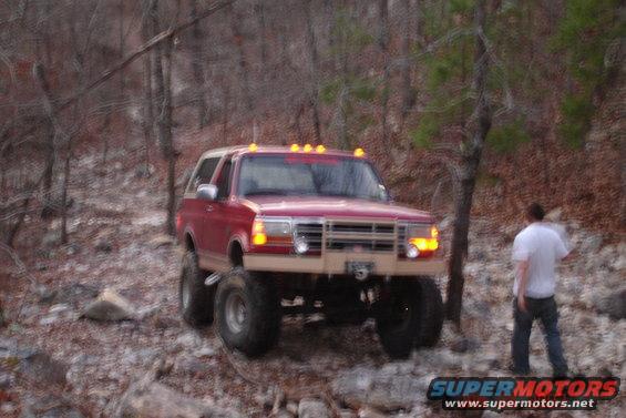 Red Ford Bronco with tan bumper and white roof, lifted with large tires, on a rocky, wooded trail