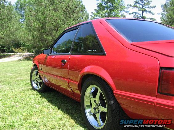Red coupe parked on grass with trees in background. Small 'Mustang' badge visible on rear quarter window