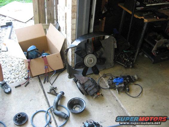 Car parts laid out on a garage floor, including a fan shroud, engine components, and a box of wires