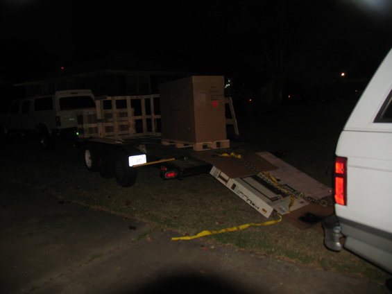 trailer is parked at night, loaded with a large box and wooden crates. A white vehicle is partially visible on the right