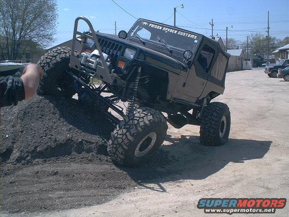 Modified Jeep Wrangler with large tires climbing a dirt mound, "PRISON SPYDER CUSTOMS" sticker on windshield