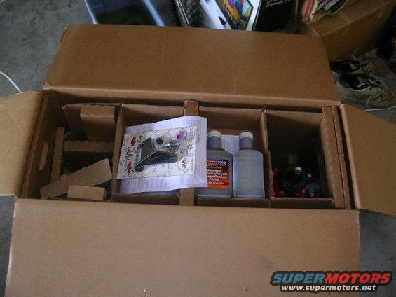 Box containing automotive parts, including a black bracket with hardware, and two bottles of Amsoil transmission fluid