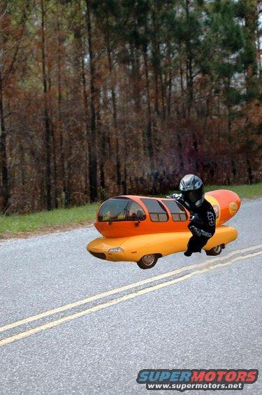 Oscar Mayer Wienermobile driving down a road with a person in motorcycle gear riding it