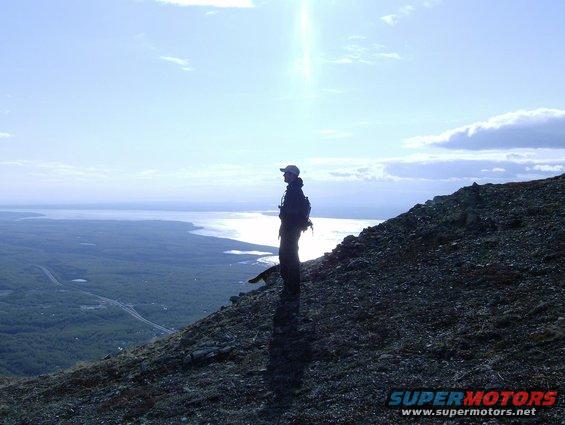 guy with a backpack on a rocky slope looking out over a large lake and some trees