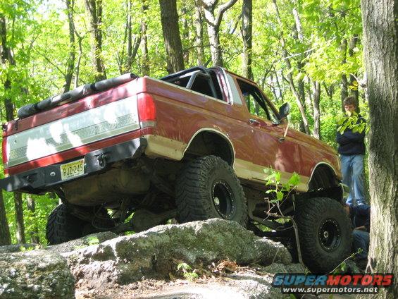 Red Ford Bronco with lifted suspension and large tires navigating rocks on a wooded trail. License plate RJZ-245 is visible