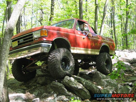 Red 1990 Ford Bronco with lifted suspension driving over rocks