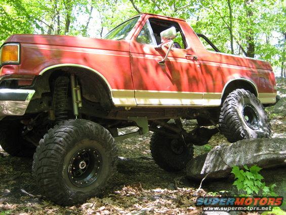 Red lifted Ford Bronco climbing over rocks on a wooded trail. It has large off-road tires and a suspension lift
