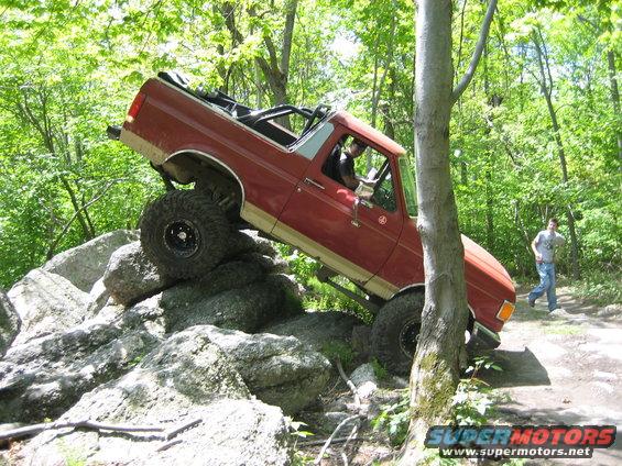 Red SUV with lifted suspension and large tires is navigating a rocky trail, with a person walking in the background