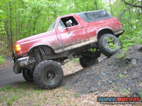 Red Ford Bronco climbing a dirt hill, lifted suspension and large tires. Black soft top
