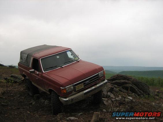Red 1990 Ford Bronco with a soft top, parked on a rocky incline with a wooded valley in the background