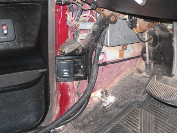 Close-up of car's driver side floor area, showing pedals, wiring, and a hood release lever