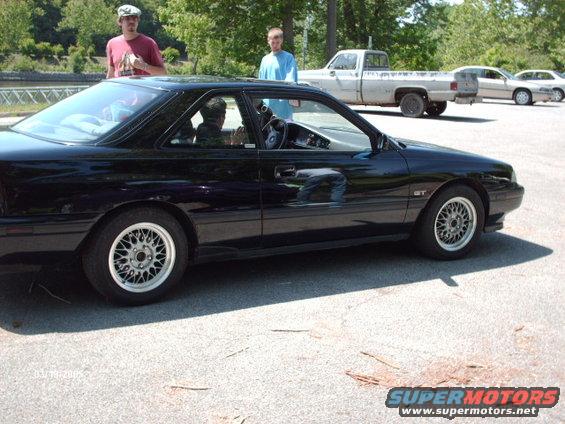 Black coupe with GT badge parked on asphalt, two people standing nearby. Pickup truck and another car in background
