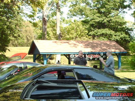 Two men stand near a pavilion with 'Mazdas' painted on it; cars are visible in the foreground