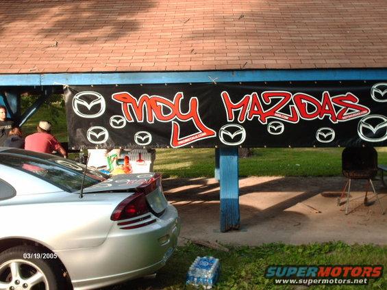 Banner says 'Indy Mazdas' with Mazda logos. A silver car is parked in front