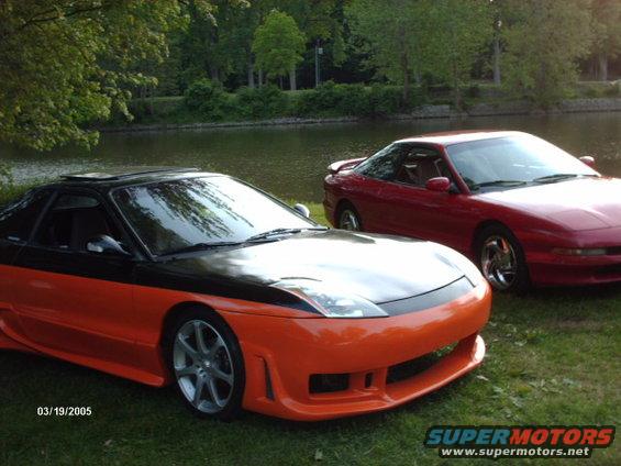 Two Ford Probe cars, one orange and black, one red, parked on grass near a body of water. Date stamp 03/19/2005
