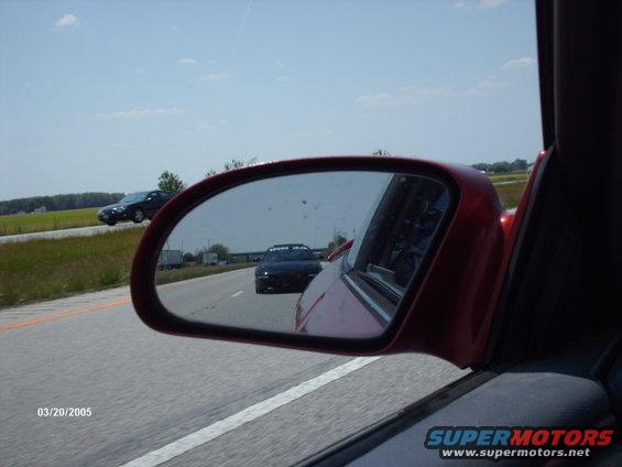 View in side mirror shows a black car ahead on highway. Another car is visible in the distance on the left