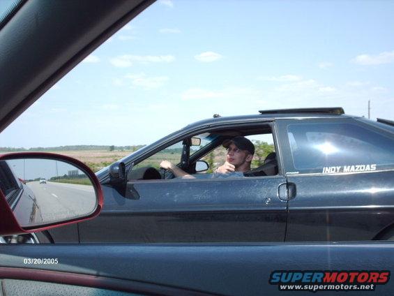 Dark coupe driving on a highway, driver wearing a baseball cap. "INDY MAZDAS" sticker on the rear window