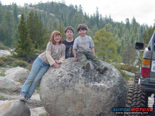 Three kids on a boulder with a lifted truck nearby. The truck has a license plate with '6' visible