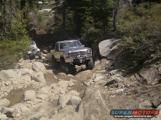 Ford Bronco driving over rocks on a trail, with a Jeep behind it