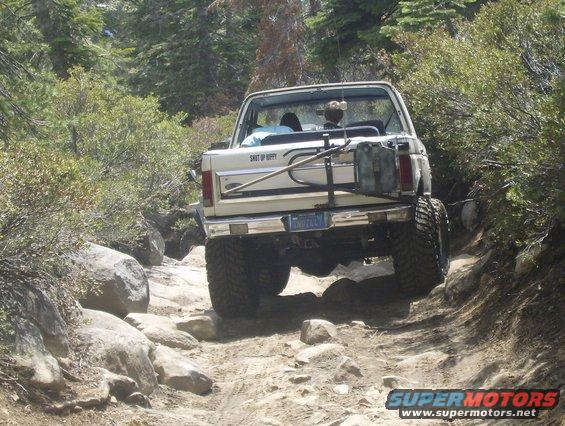 White Ford Bronco on a rocky trail, rear view. It has a spare tire carrier with a gas can