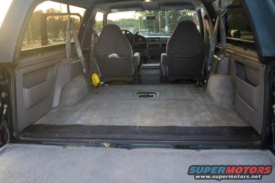 Interior of a dark blue Ford Bronco, showing the rear cargo area and front seats. The carpeted floor is visible, along with door panels and seatbelts