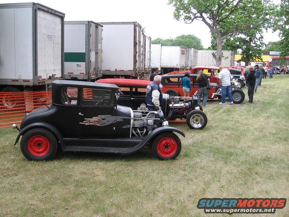 Several hot rod cars parked on grass, with trailers in the background. One black car in front has red wheels and flame decals