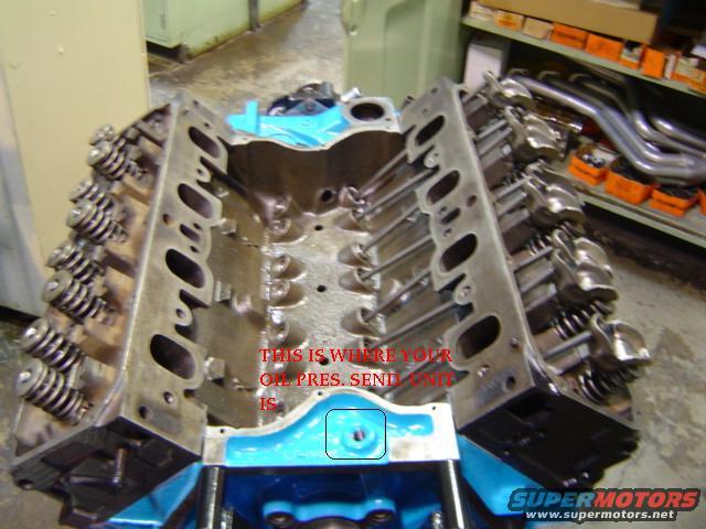 Engine block with blue painted components and visible valve train components. Text overlay indicates oil pressure sensor location